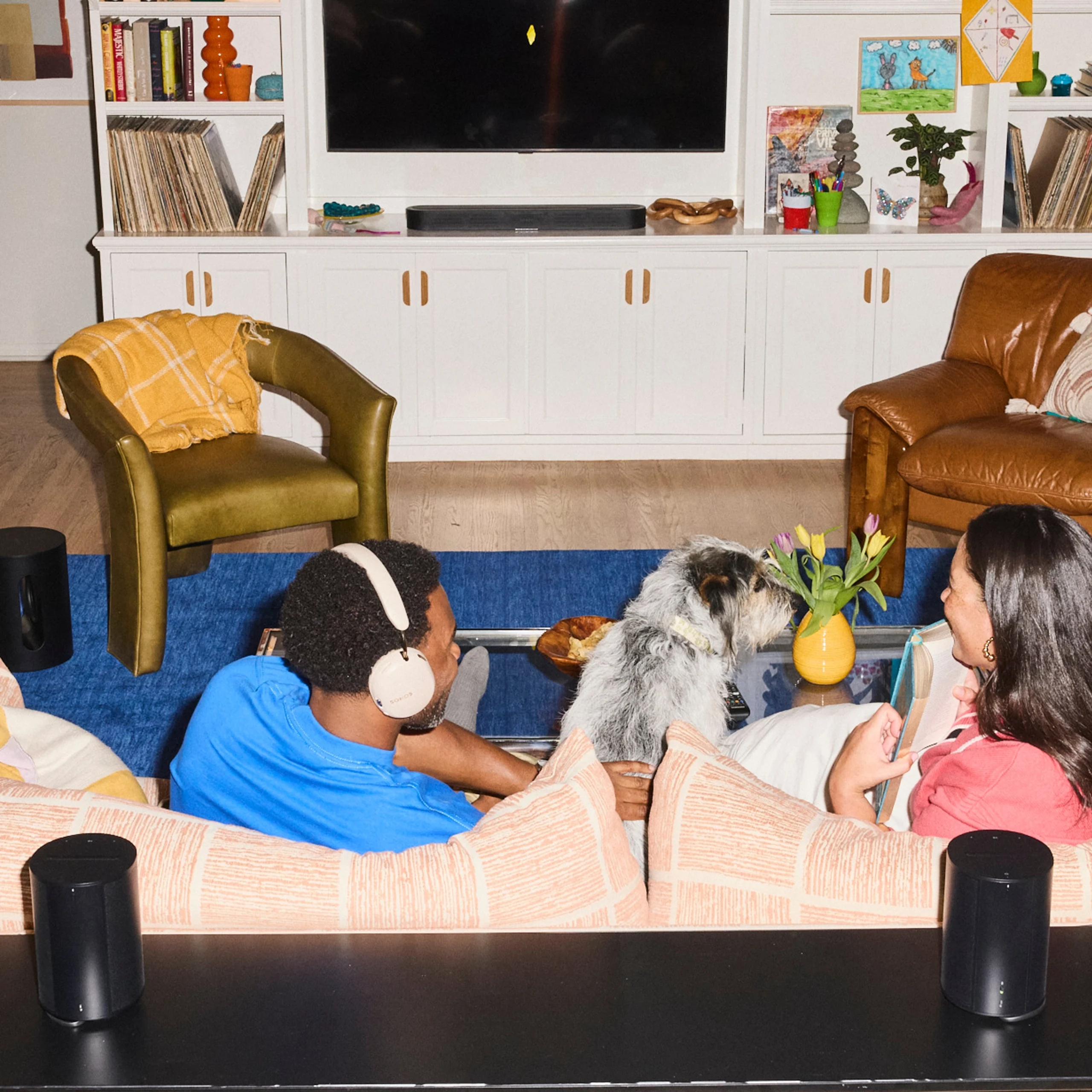 A cozy home theater scene with two people enjoying music with a dog, surrounded by stylish decor and a TV.
