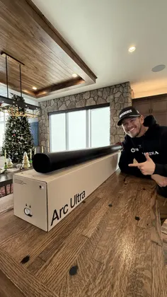 A person points at the Arc Ultra speaker on a wooden table, with a festive tree and stone wall in the background.